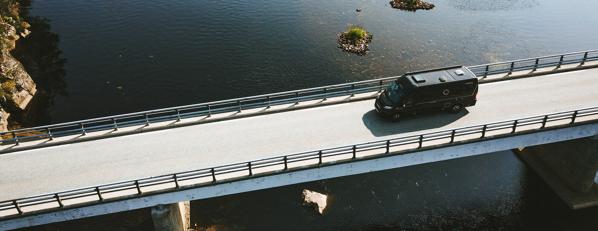 Fahraufnahme aus der Vogelperspektive von einem Malibu Van auf einer Brücke über einem See