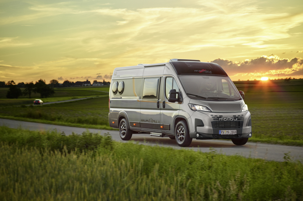 Malibu Campervan auf der Straße, umgeben von Wiesen im Hintergrund der Sonnenaufgang.