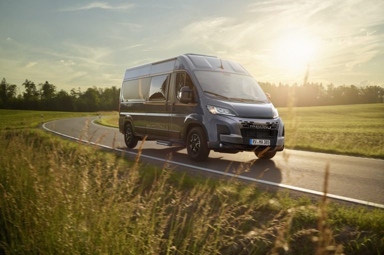 Grauer Malibu Campervan auf Landstraße im Hintergrund Wald.