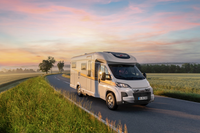 Teilintegriertes Malibu Reisemobil auf der Landstraße umgeben von Wiesen. Im Hintergrund Sonnenaufgang.