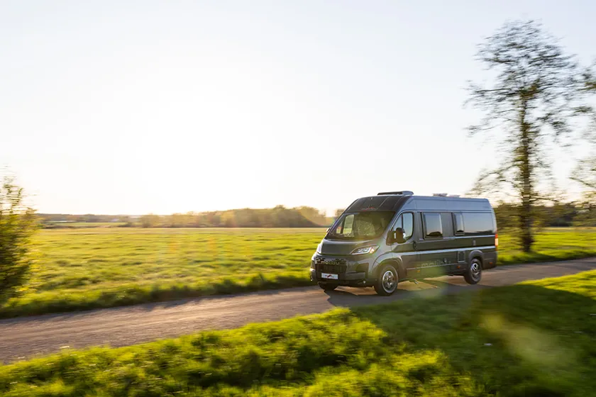 Malibu Camper Van fährt bei Sonnenuntergang durch grüne Landschaft auf einer Landstraße