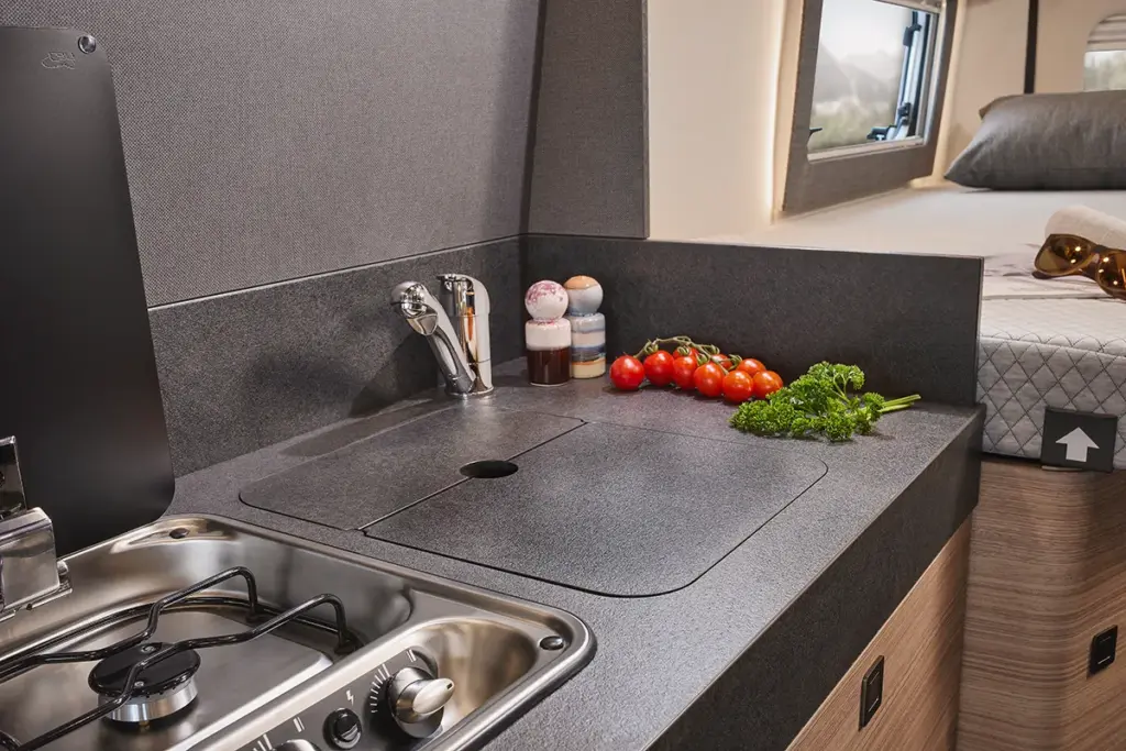 Une cuisine compacte de camping-car équipée d'un évier, d'une cuisinière à gaz et de légumes frais, comme des tomates et du persil, posés sur le plan de travail. On aperçoit à l'arrière-plan un lit avec des oreillers et des baies.