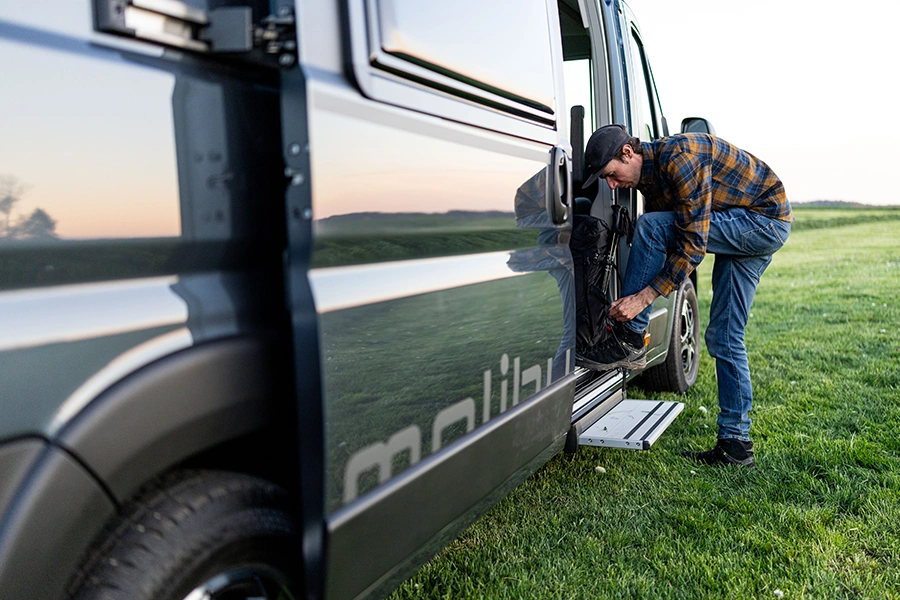 Camper steigt am Malibu Relax Van auf einer Wiese aus und schnürt seine Wanderschuhe auf dem Einstieg des Fahrzeug