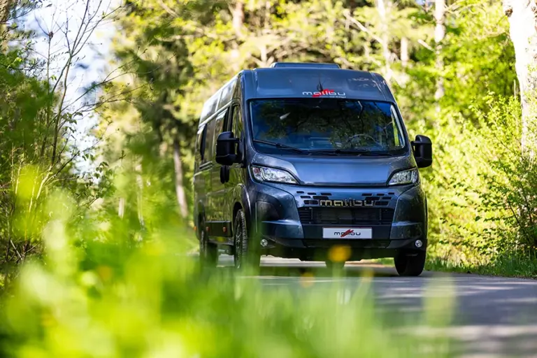 Malibu Relax Camper Van auf einer sonnigen Landstraße zwischen Wiesen und Wald
