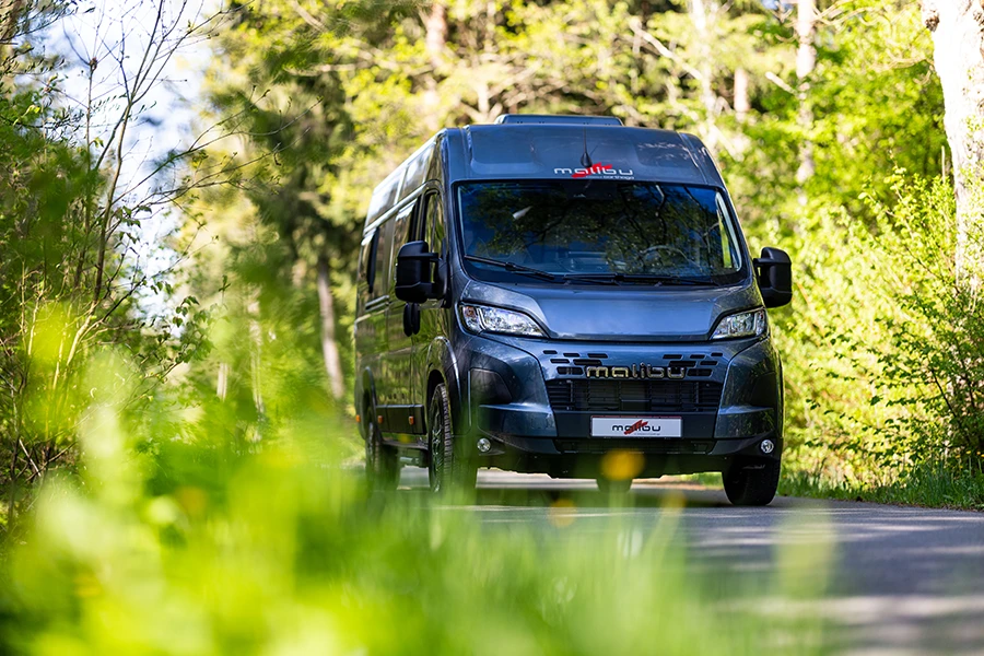 Malibu Relax Camper Van auf einer sonnigen Landstraße zwischen Wiesen und Wald