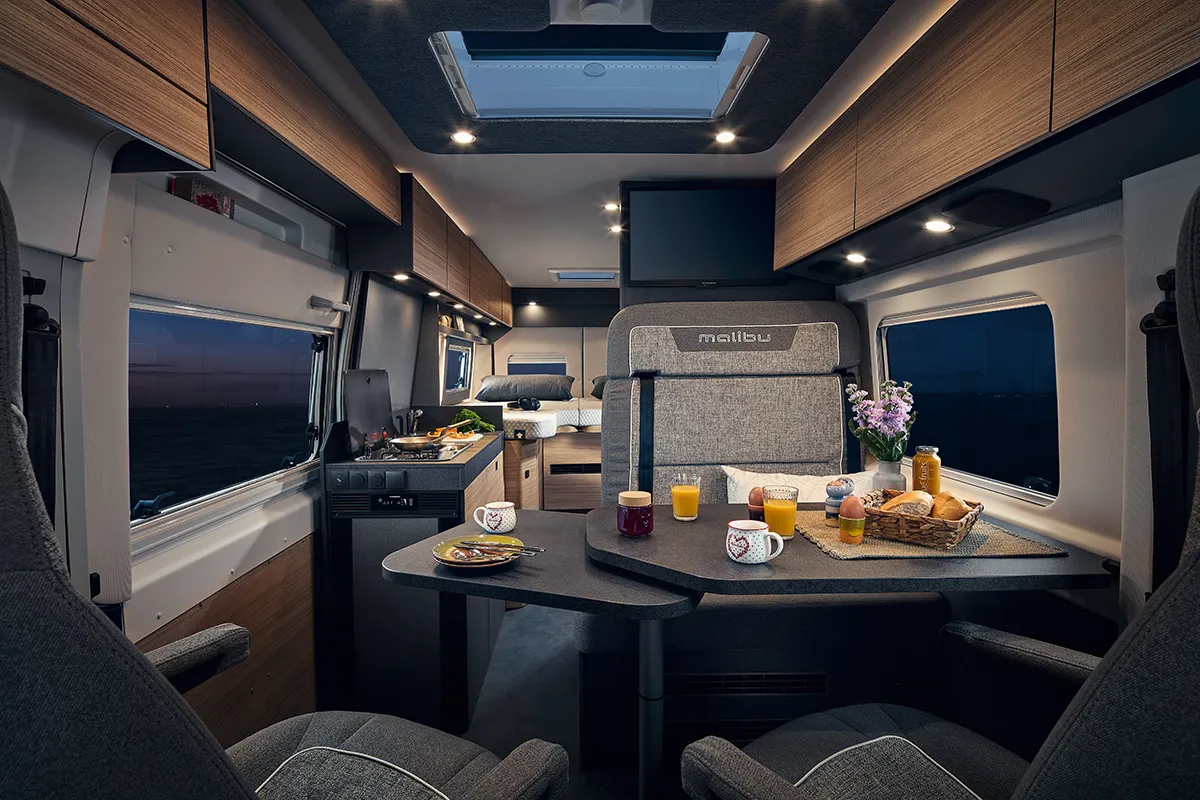 Gemütlicher Wohn- und Essbereich im Malibu Relax Camper Van mit gedecktem Frühstückstisch, stilvoller Innenbeleuchtung, modernem Interieur in Holzoptik