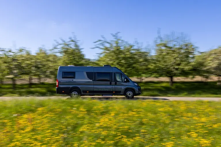 Malibu Relax Camper Van fährt im Frühling durch blühende Landschaft mit gelben Blumen und Obstbäumen