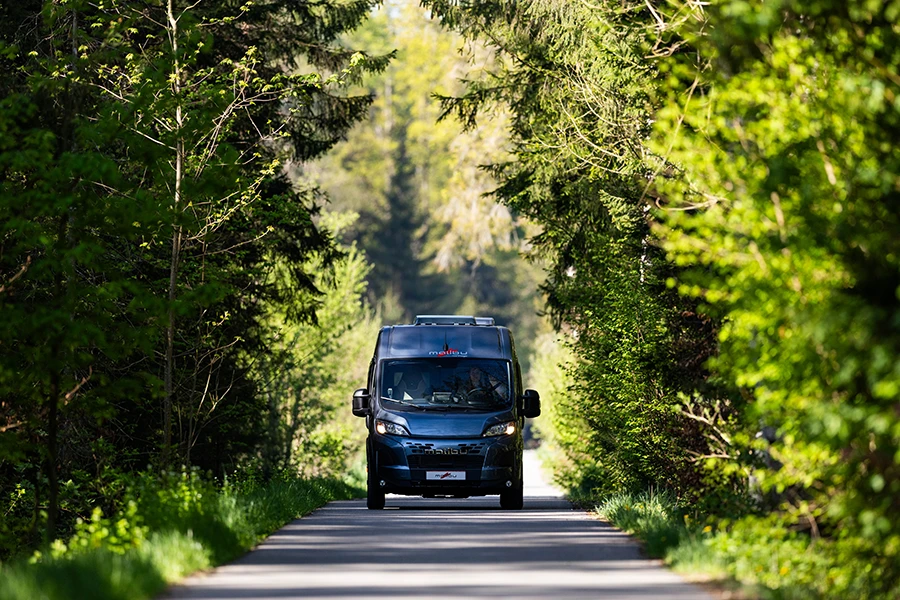 Malibu Relax Camper Van fährt durch eine von Bäumen gesäumte Waldstraße, Blick auf das Fahrzeug von vorne
