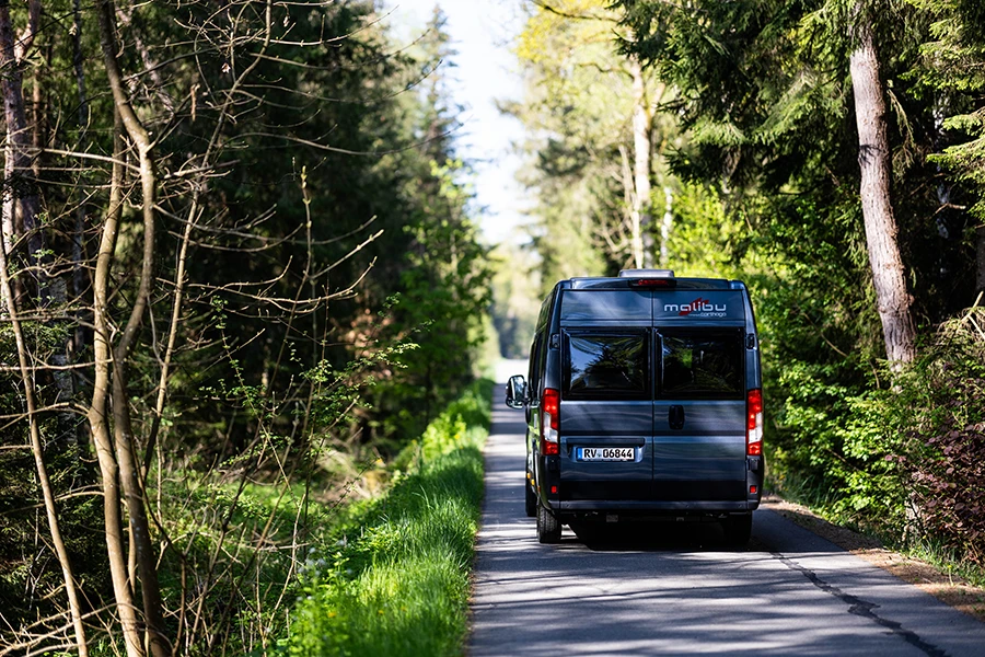 Malibu Relax Camper Van fährt durch eine von Bäumen gesäumte Waldstraße, Blick auf das Fahrzeug von hinten