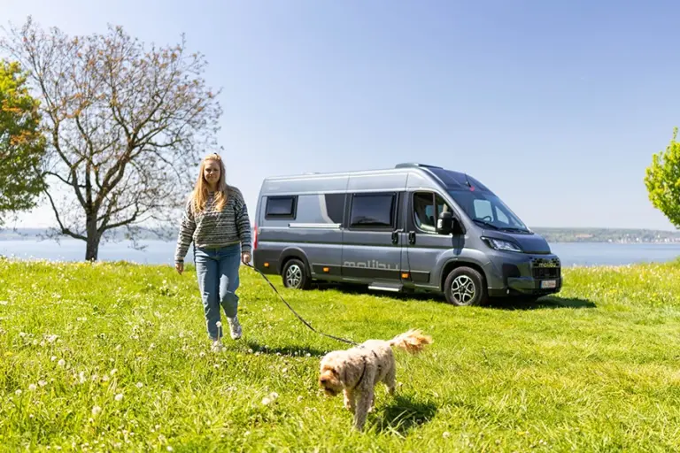 Frau mit Hund spaziert vor einem geparkten Malibu Relax Van am Seeufer auf einer grünen Wiese entlang