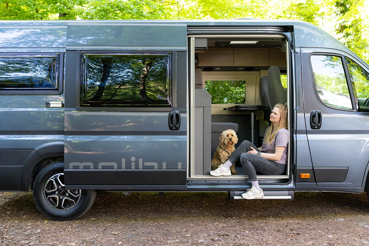 Junge Frau sitzt mit ihrem Hund in der geöffneten Seitentür eines Malibu Relax Camper Vans – geparkt im Wald, umgeben von Natur und Tageslicht, ideal für Reisen mit Hund.