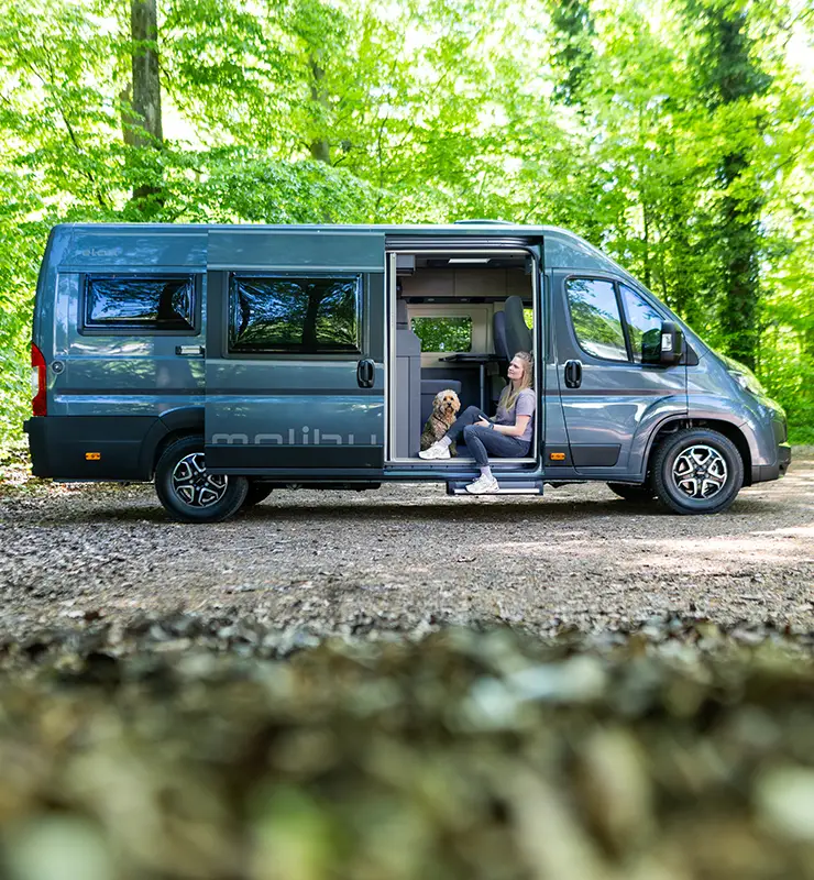 Frau entspannt mit Hund im geöffneten Malibu Camper Van im grünen Wald