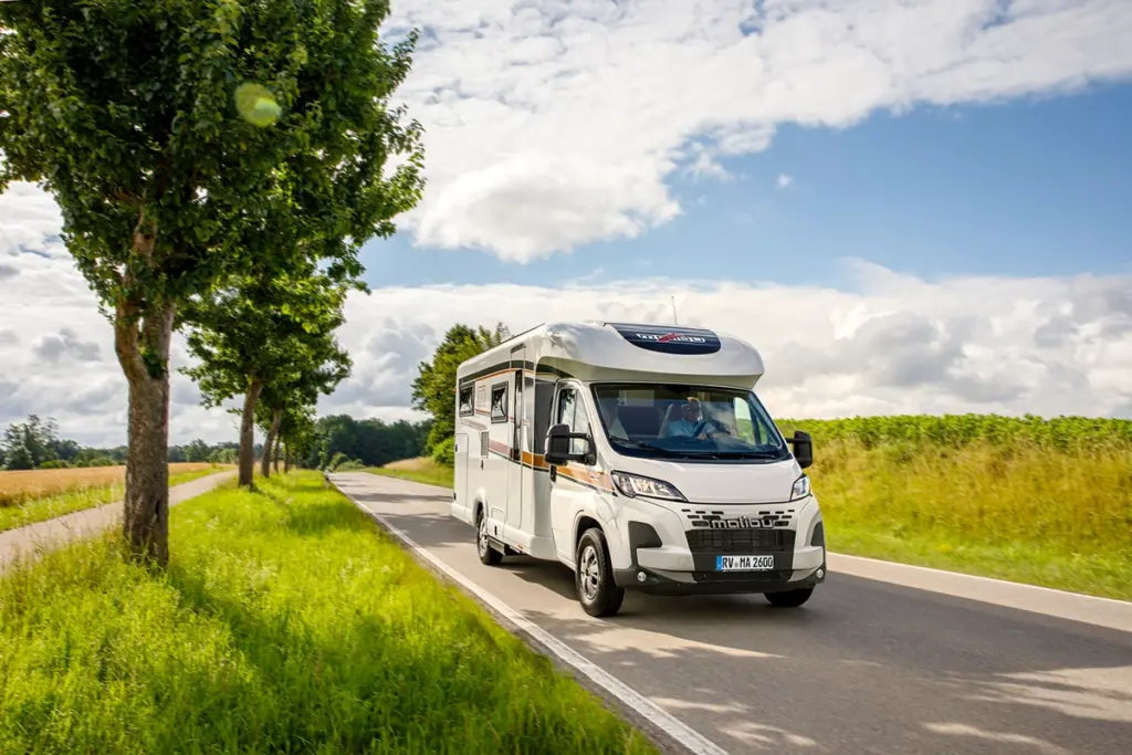 Ein weißes Wohnmobil fährt unter teilweise bewölktem Himmel eine von grünem Gras und Bäumen gesäumte Landstraße entlang.