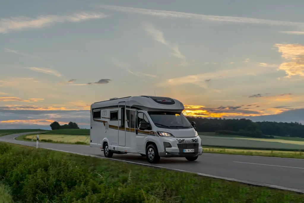 Ein weißes Wohnmobil fährt bei Sonnenuntergang auf einer Landstraße, mit grünen Feldern auf beiden Seiten und einem bunten Himmel mit Wolken im Hintergrund.