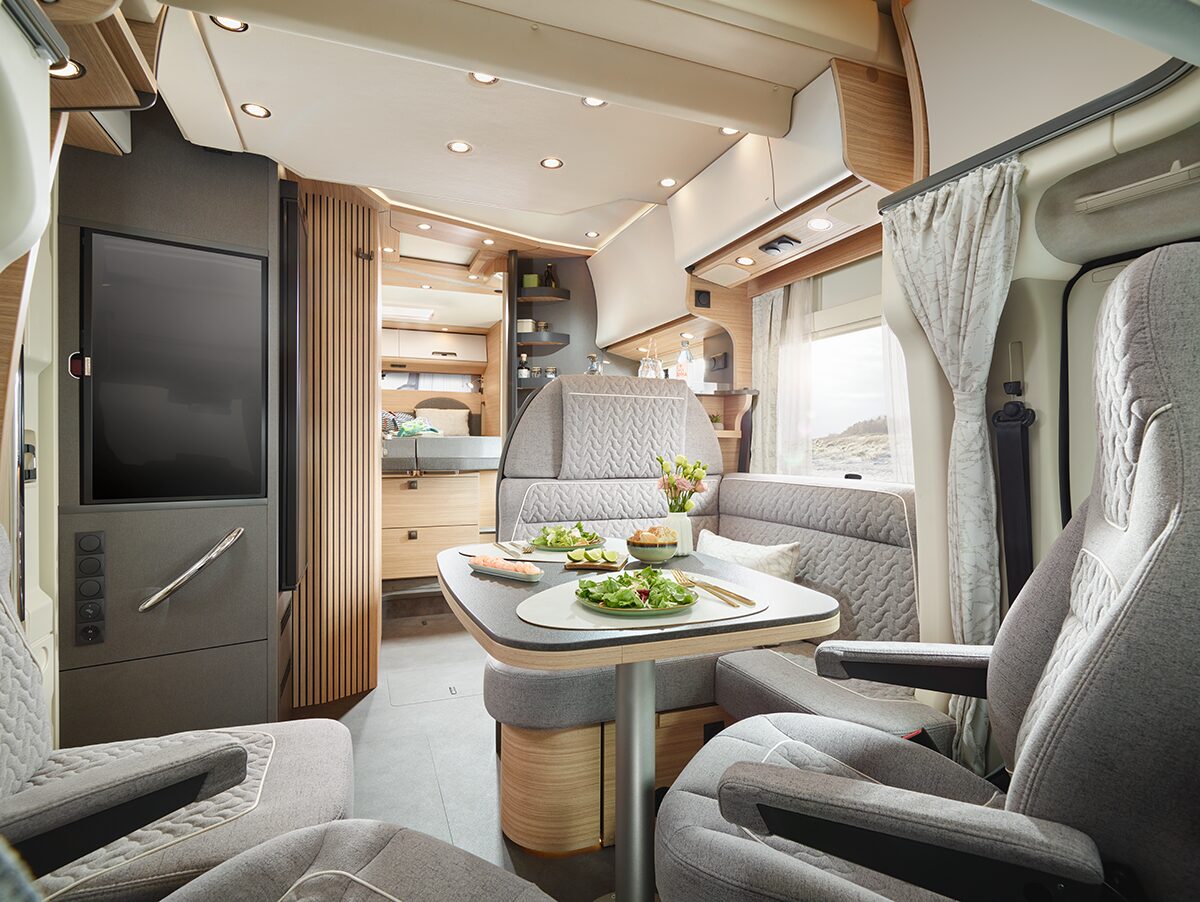 Modernes Wohnmobil-Interieur mit grauen gepolsterten Sitzen und einem Esstisch für vier Personen mit Salaten. Der Raum ist gut beleuchtet, mit Holzschränken, Regalen und einem Fenster mit Vorhängen, die eine gemütliche und organisierte Atmosphäre schaffen.