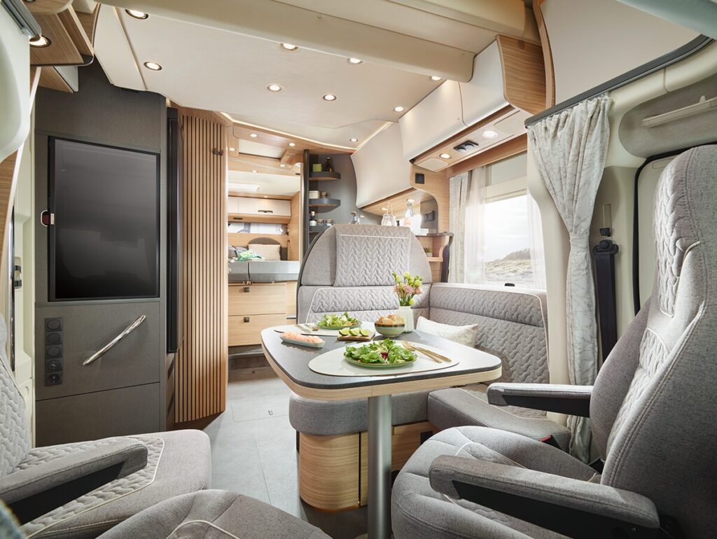 Modernes Wohnmobil-Interieur mit grauen gepolsterten Sitzen und einem Esstisch für vier Personen mit Salaten. Der Raum ist gut beleuchtet, mit Holzschränken, Regalen und einem Fenster mit Vorhängen, die eine gemütliche und organisierte Atmosphäre schaffen.