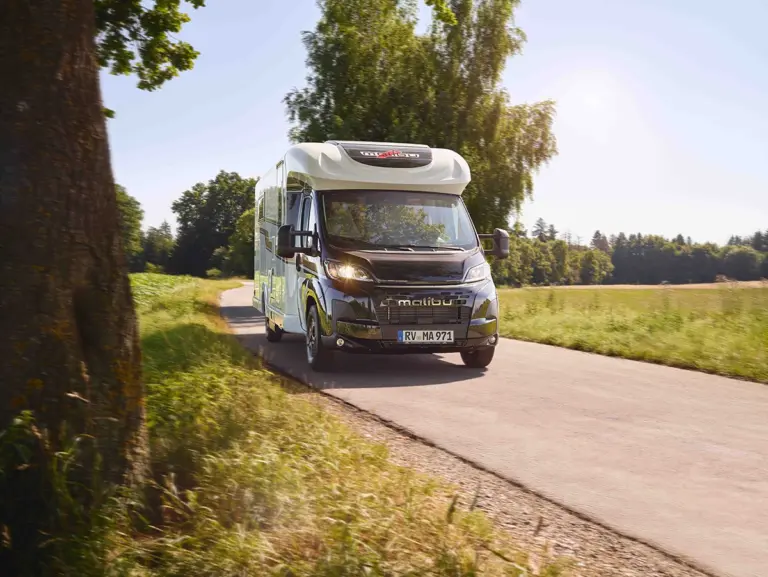 Teilintegriertes Malibu Reisemobil in weiß mit schwarzer Front fährt auf einer Straße mit Sonnenlicht und Bäumen, Frontansicht