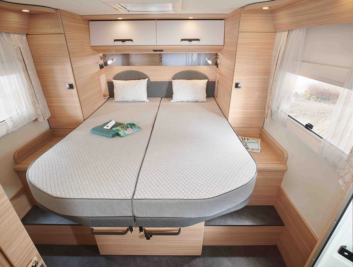 Zwei Einzelbetten mit grauer Bettwäsche stehen in einem gemütlichen Wohnmobil-Schlafzimmer nebeneinander und verschmelzen zu einem großen Schlafbereich. Der Raum ist mit Holzvertäfelung, Hängeschränken und Fenstern mit Vorhängen ausgestattet.