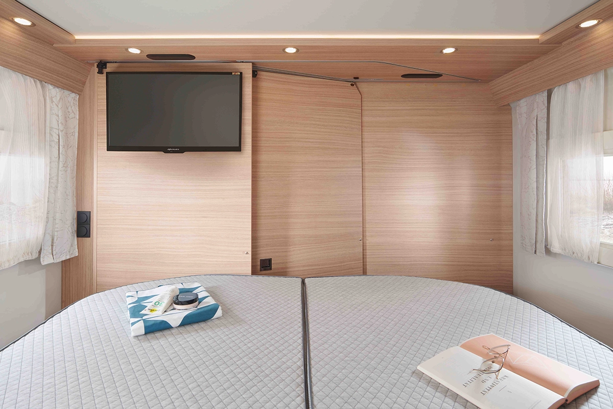 Ein modernes Wohnmobil-Schlafzimmer mit hellen Holzwänden, zwei Betten mit hellgrauen Steppdecken, einem Flachbildfernseher an der Wand und Fenstern mit durchsichtigen Vorhängen. Ein aufgeschlagenes Buch und ein Tablett mit einer Tasse und Essen stehen auf den Betten.