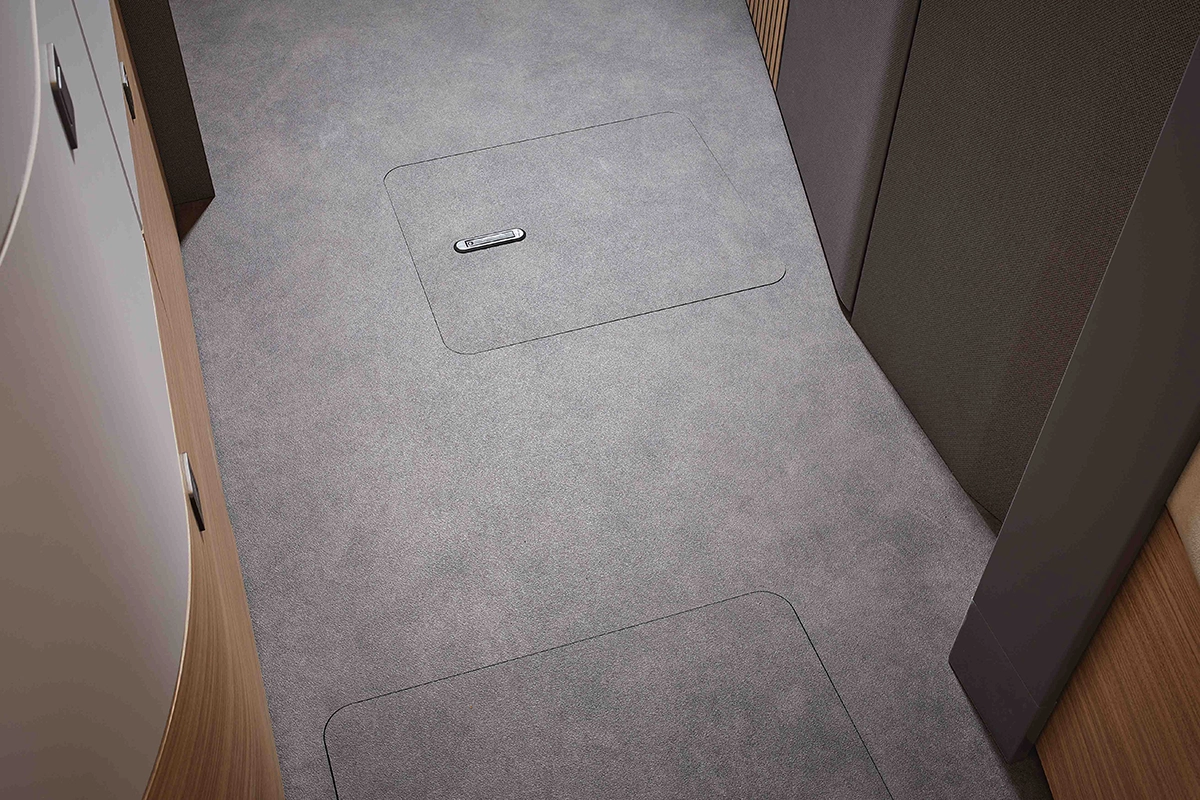 Nahaufnahme eines hellgrauen Teppichbodens in einem Flur mit zwei rechteckigen Zugangspaneelen mit einem versenkten Griff an einem Paneel. Wände mit Holz- und Stoffverkleidungen säumen die Seiten.