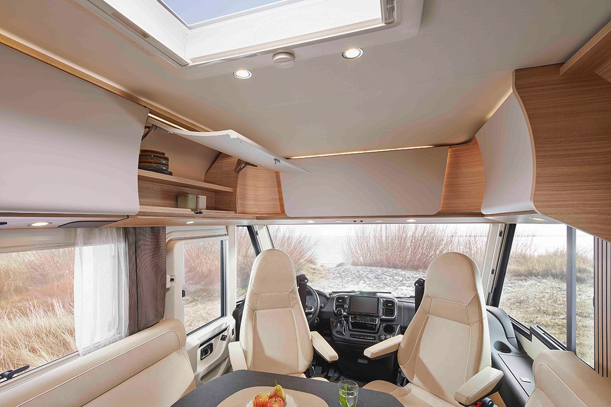 Das Innere eines modernen Wohnmobils zeigt zwei cremefarbene Kapitänssessel, ein Lenkrad, einen runden Tisch mit Obst, eingebaute Holzschränke, große Fenster und ein Dachfenster, mit Blick ins Freie im Hintergrund.