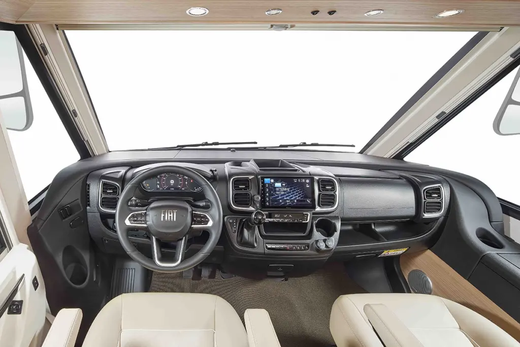 Interiörvy av en modern cockpit i en husbil med Fiat-ratt, reglage på instrumentbrädan, pekskärm och säten i beige läder. Designen är ren och rymlig med stora fönster.