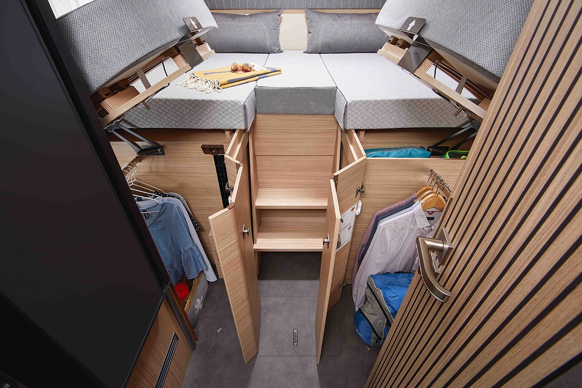 Ein kompaktes Schlafzimmer mit einem erhöhten Bett, unter dem sich Ablagefächer befinden. Die Kleidung hängt ordentlich in den Schränken auf beiden Seiten, und in den Regalen unter dem Bett finden verschiedene Gegenstände Platz. Helles Holz und Grautöne schaffen einen modernen, organisierten Raum.