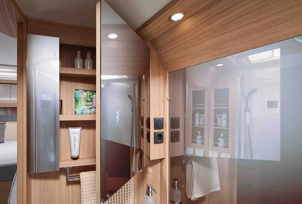 Ein modernes Wohnmobil-Badezimmer mit Holzverkleidung, Spiegelschränken, Regalen mit Toilettenartikeln und einem ordentlich gefalteten Handtuch. Spiegelungen zeigen eine Dusche, einen Teil des Wohnbereichs und Deckenleuchten.