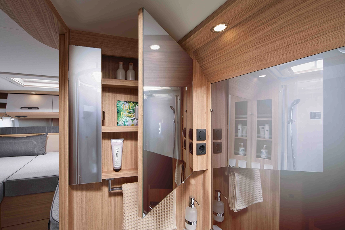 Ein modernes Wohnmobil-Badezimmer mit Holzverkleidung, Spiegelschränken, Regalen mit Toilettenartikeln und einem ordentlich gefalteten Handtuch. Spiegelungen zeigen eine Dusche, einen Teil des Wohnbereichs und Deckenleuchten.