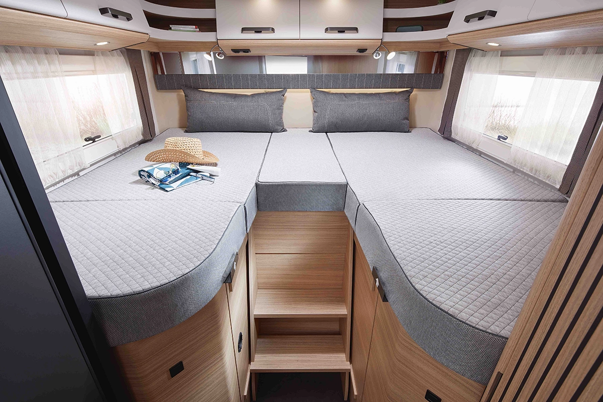 Modernes Wohnmobil-Interieur mit Einzelbetten, grauem Bettzeug, Holzschränken und großen Fenstern mit Vorhängen. Auf einem Bett liegen ein Hut, ein Buch und eine Sonnenbrille, und zwischen den Betten führen Stufen nach oben.