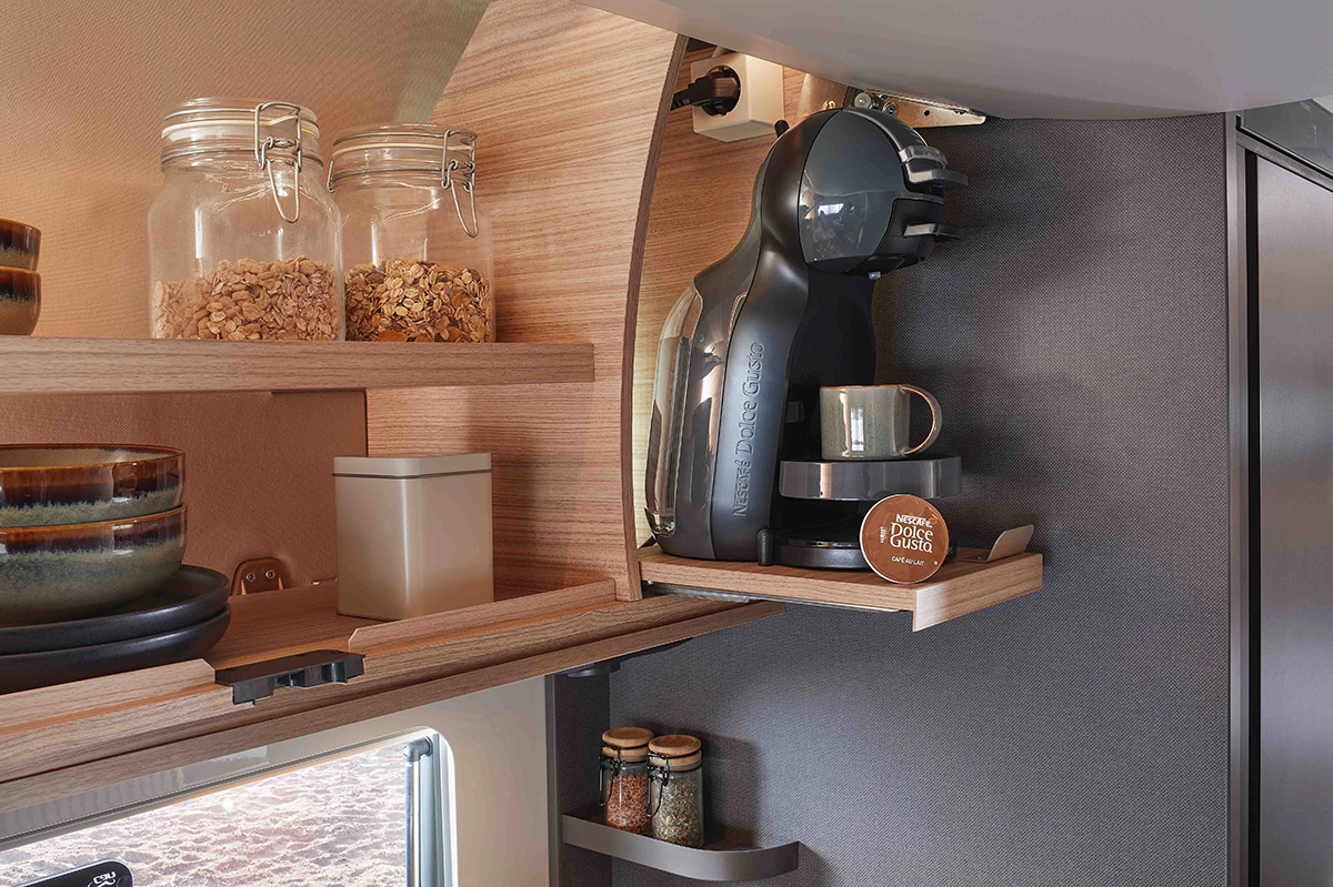 Ein kompaktes Küchenregal beherbergt eine Kaffeemaschine mit einer Tasse, Kaffeepads und Gläsern mit Müsli darüber. In den benachbarten Regalen stehen gestapelte Schüsseln und Gläser mit Gewürzen. Die Einrichtung ist ordentlich und modern mit Holzakzenten.
