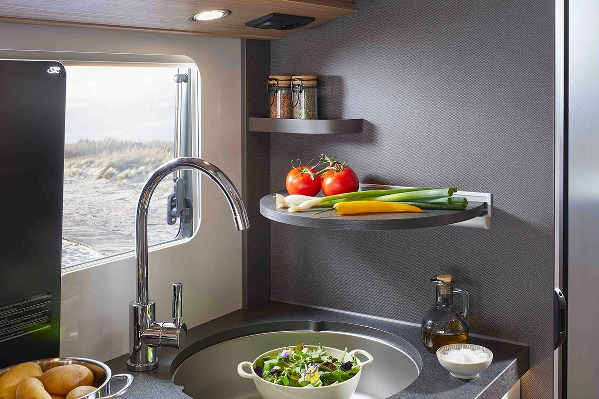 Moderne Wohnmobilküche mit runder Spüle, Salatschüssel, frischem Gemüse auf einem Regal, Gewürzen, Olivenöl und einem malerischen Sandstrand vor dem Fenster.