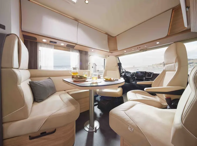 Modernes Wohnmobil-Interieur mit cremefarbenen Ledersitzen, einem kleinen Esstisch für zwei Personen und großen Fenstern, die natürliches Licht hereinlassen. Der Fahrerbereich ist einsehbar, und die Einrichtung ist in hellem Holz und neutralen Tönen gehalten.