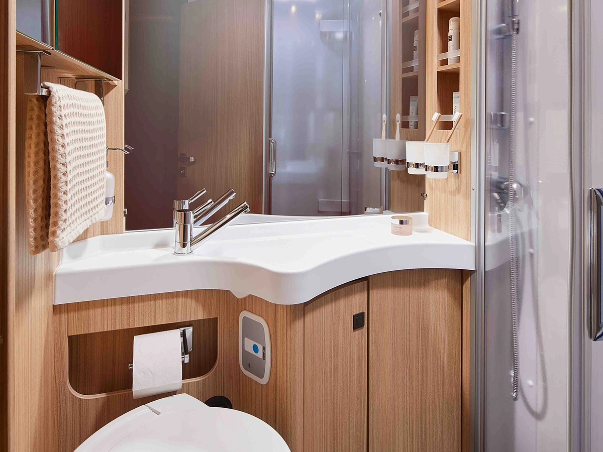 Modernes Badezimmer mit hellen Holzschränken, einer weißen Arbeitsplatte und einem Waschbecken, einem Handtuch auf einem Ständer, Toilettenpapier und Regalen mit Toilettenartikeln. Auf der rechten Seite befindet sich eine Duschkabine aus Glas.