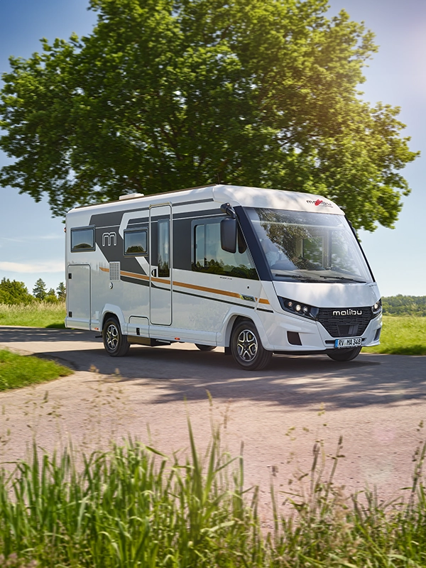 2026 MalibuReisemobil RM2 I430KB LE K Lightweight 3.5t FIAT Higlights Exterieur Ein weißes Malibu-Wohnmobil fährt auf einer sonnigen Landstraße, umgeben von grünem Gras, Bäumen und fernen Hügeln unter einem klaren blauen Himmel.