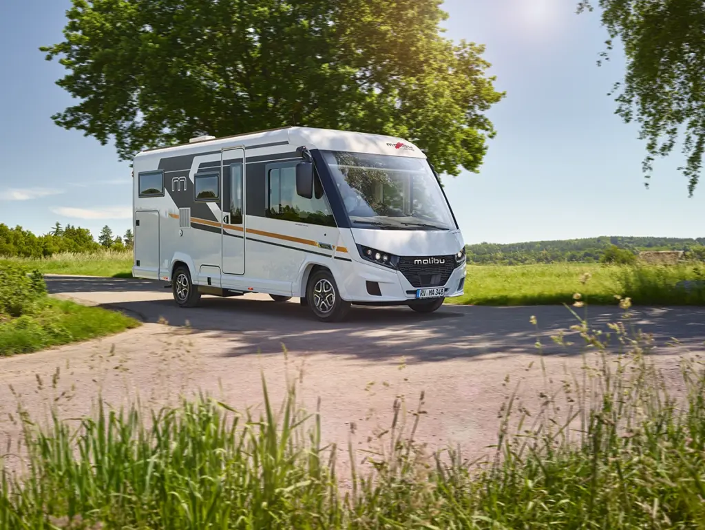 Ein weißes Wohnmobil fährt an einem sonnigen Tag unter strahlend blauem Himmel eine Landstraße entlang, die von grünem Gras und Bäumen gesäumt ist.