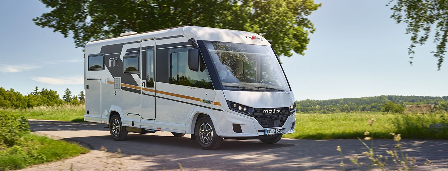 2026 MalibuReisemobil RM2 I430KB LE K Lightweight 3.5t FIAT Higlights Exterieur Ein weißes Malibu-Wohnmobil fährt auf einer sonnigen Landstraße, umgeben von grünem Gras, Bäumen und fernen Hügeln unter einem klaren blauen Himmel.