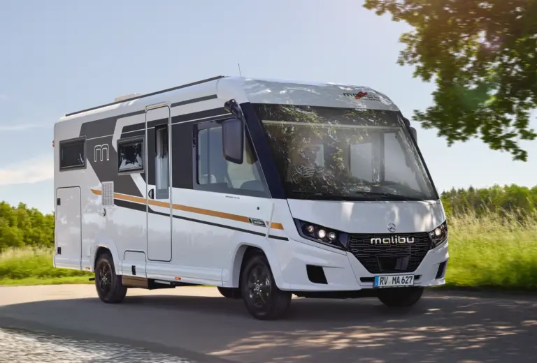 Vollintegriertes Malibu Reisemobil in weiß fährt auf einer Straße mit Sonnenlicht, Bäumen und blauem Himmel
