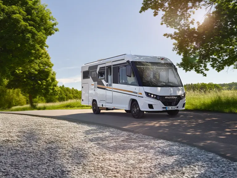 Ein weißes Malibu-Wohnmobil fährt auf einer sonnigen, von Bäumen gesäumten Landstraße, mit grünem Gras und Bäumen auf beiden Seiten und Sonnenlicht, das durch die Äste fällt.