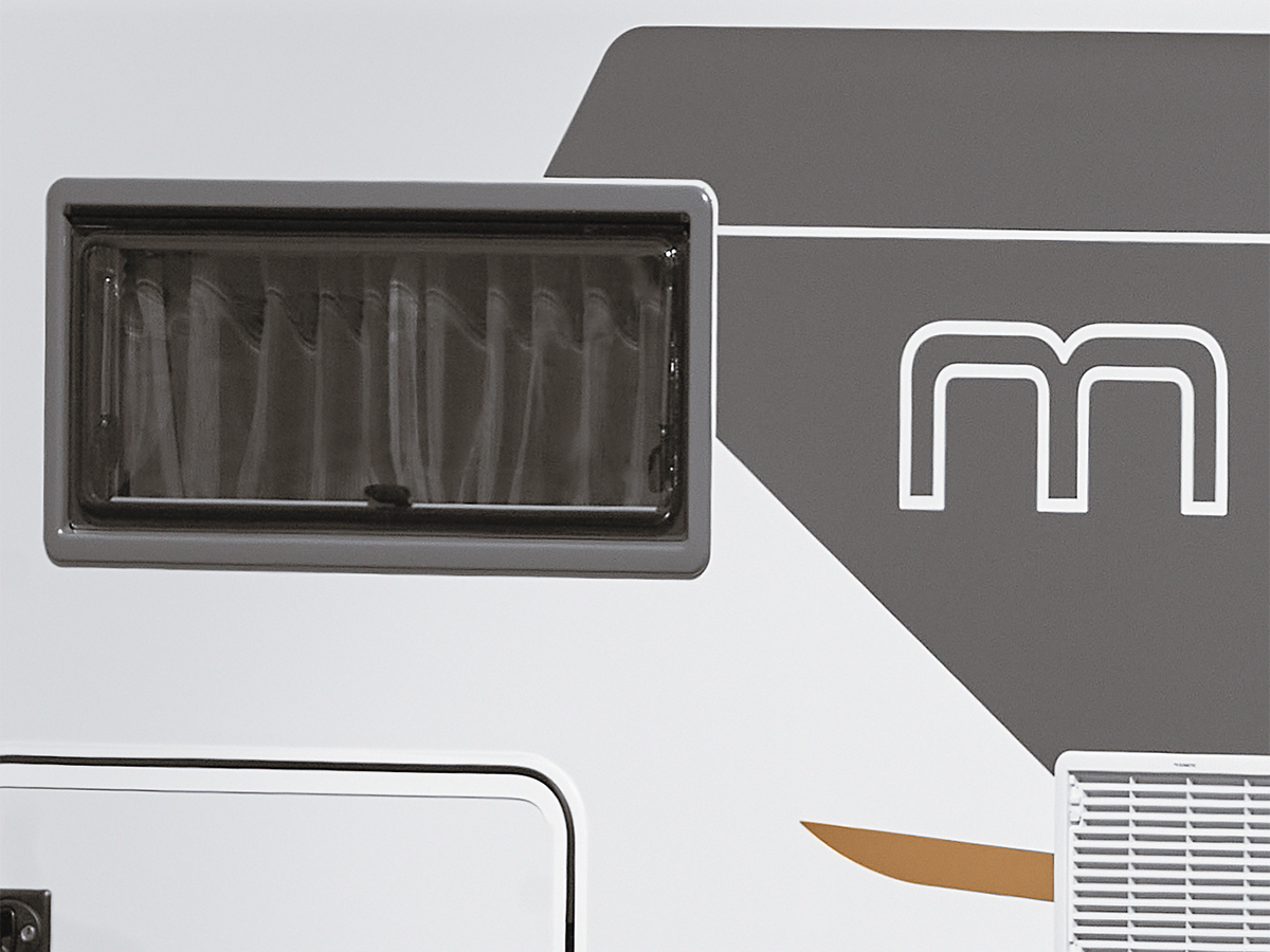 Nahaufnahme der Seite eines weiß-grauen Fahrzeugs, möglicherweise eines Wohnmobils, mit einem Fenster mit Vorhängen, einer stilisierten m-Grafik und einer kleinen Lüftungsöffnung oder -platte.