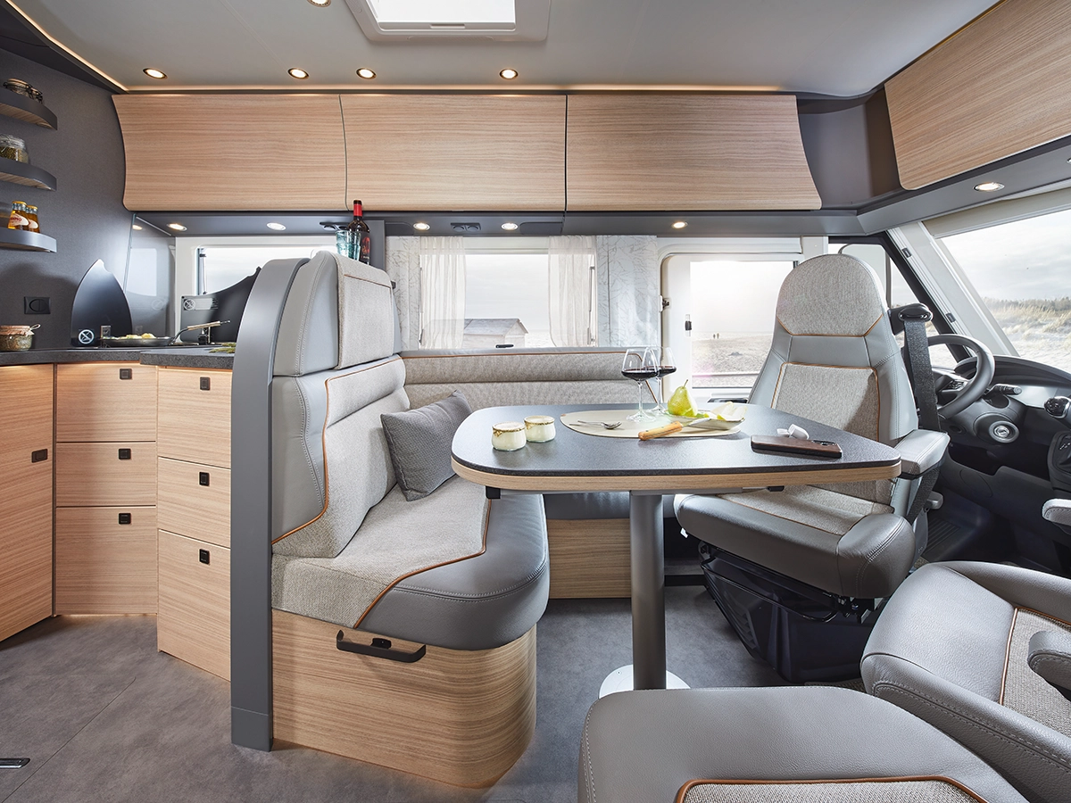 Modernes Wohnmobil-Interieur mit hellen Holzschränken, gepolsterten Sitzen um einen Esstisch und einem zum Wohnbereich hin ausgerichteten Fahrersitz. Auf dem Tisch stehen Teller mit aufgeschnittenem Obst. Durch die Fenster strömt natürliches Licht herein.
