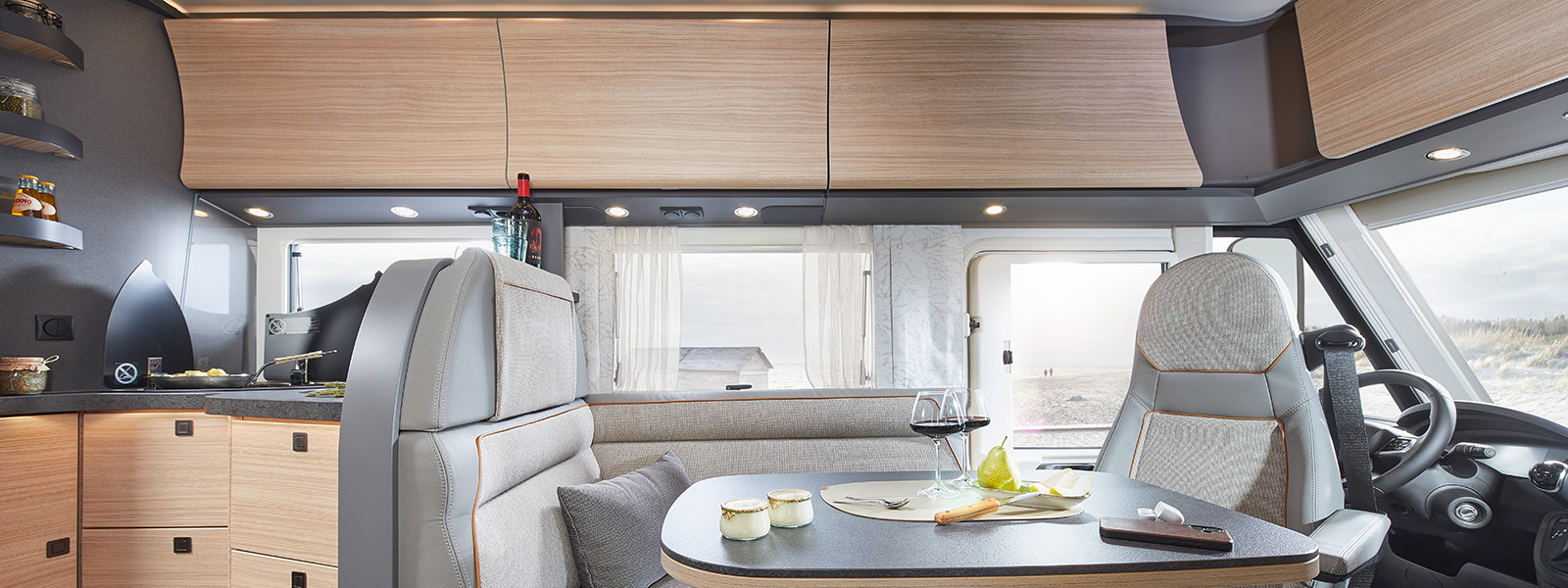 Modernes Wohnmobil-Interieur mit hellen Holzschränken, einer kleinen Küchenzeile und einem Esstisch mit Wein, Käse und Obst. Große Fenster bieten einen Blick auf den Sandstrand und den Himmel draußen.