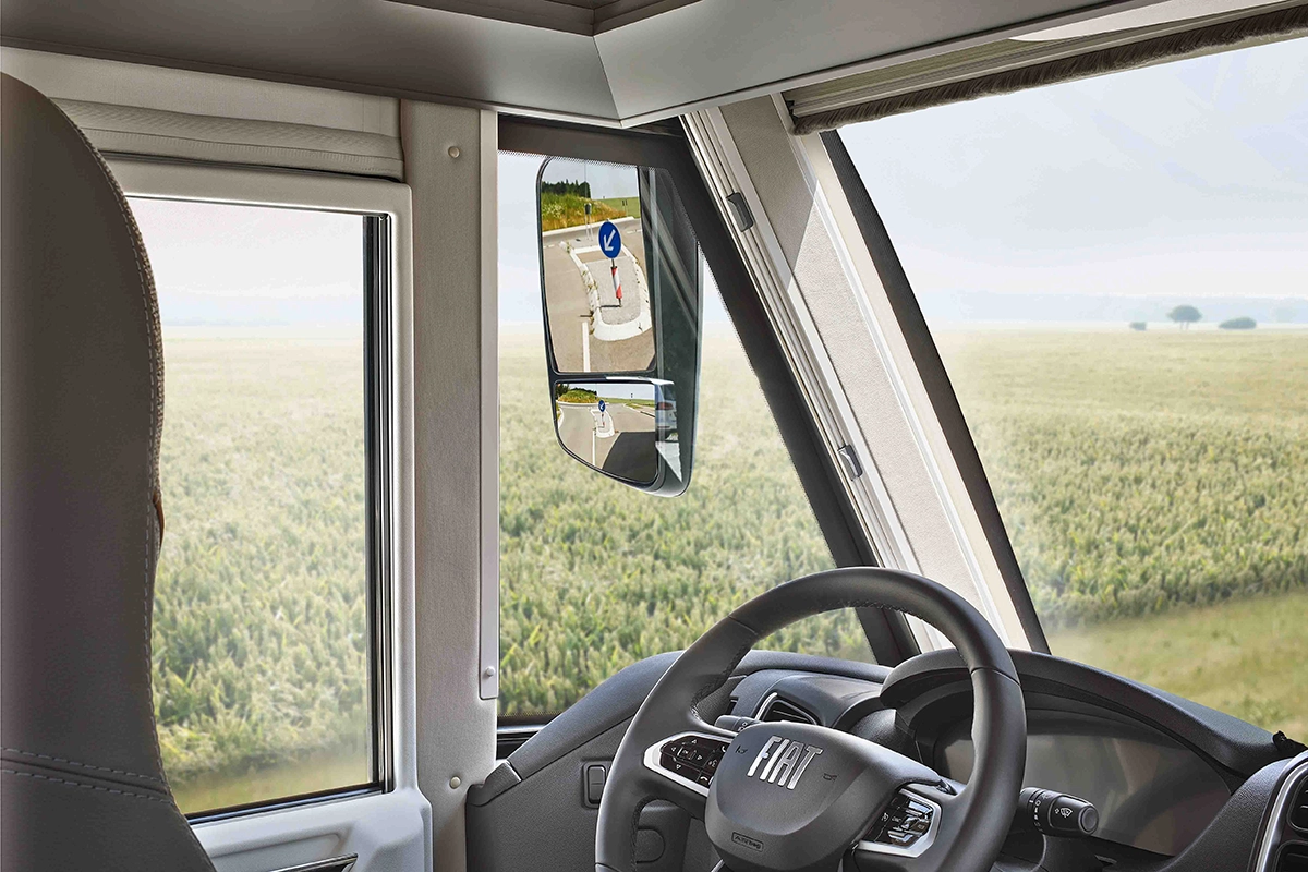 Blick vom Fahrersitz eines Fiat-Fahrzeugs auf das Lenkrad, das Seitenfenster und den Rückspiegel, in dem sich ein Straßenschild spiegelt, vor einem grünen Feld und einem bewölkten Himmel.