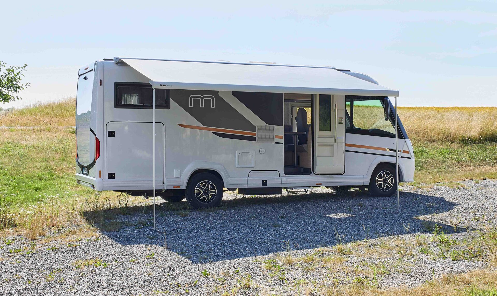 Ein weiß-graues Wohnmobil mit schwarzen und orangefarbenen Akzenten ist auf einer Kiesfläche neben einer Wiese unter freiem Himmel geparkt. Ein Sonnensegel spendet Schatten neben dem Fahrzeug.