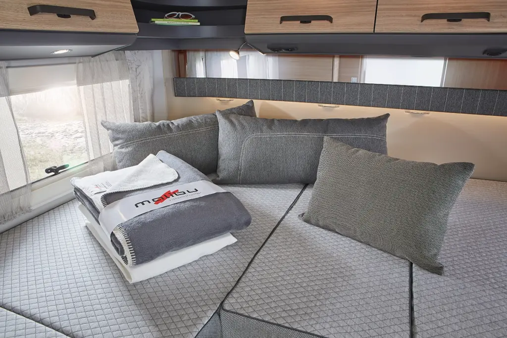 Ein gemütliches Wohnmobilbett mit grauen Kissen und Polstern, ordentlich gefalteten Handtüchern und einer Decke obenauf, unter Dachschränken und neben Fenstern mit durchsichtigen Vorhängen.