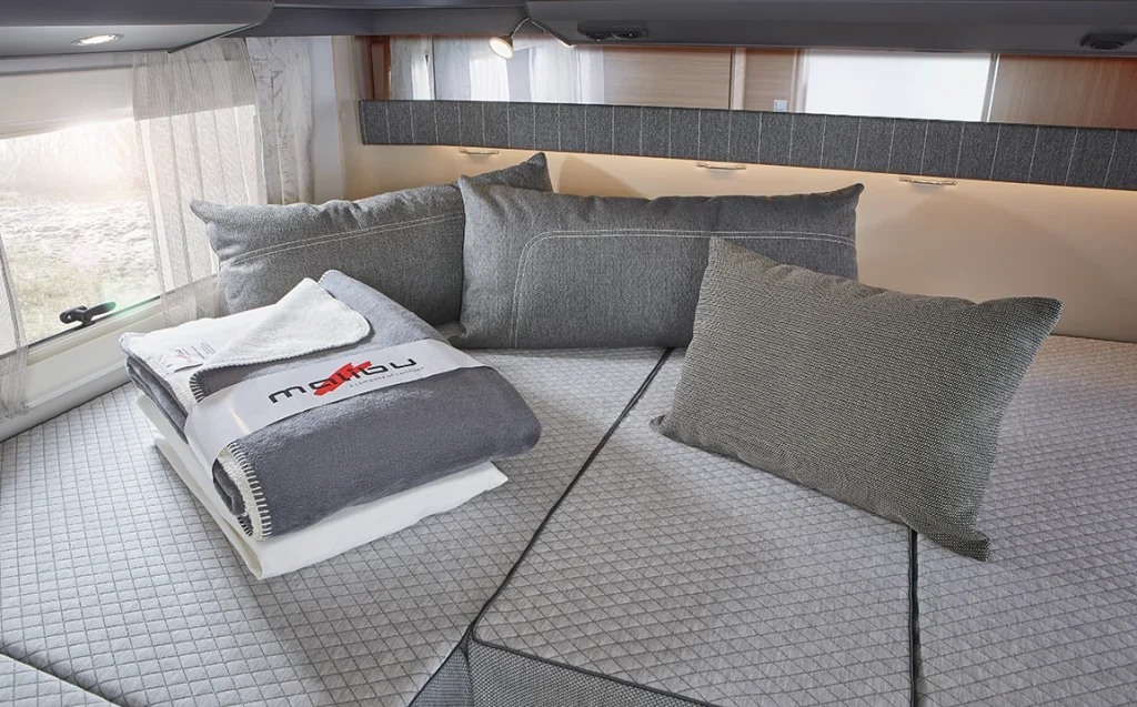 Ein gemütliches Wohnmobilbett mit grauen Kissen und Polstern, ordentlich gefalteten Handtüchern und einer Decke obenauf, unter Dachschränken und neben Fenstern mit durchsichtigen Vorhängen.
