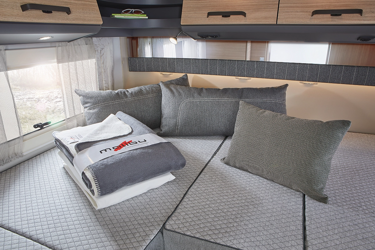 Ein gemütliches Wohnmobilbett mit grauen Kissen und Polstern, ordentlich gefalteten Handtüchern und einer Decke obenauf, unter Dachschränken und neben Fenstern mit durchsichtigen Vorhängen.