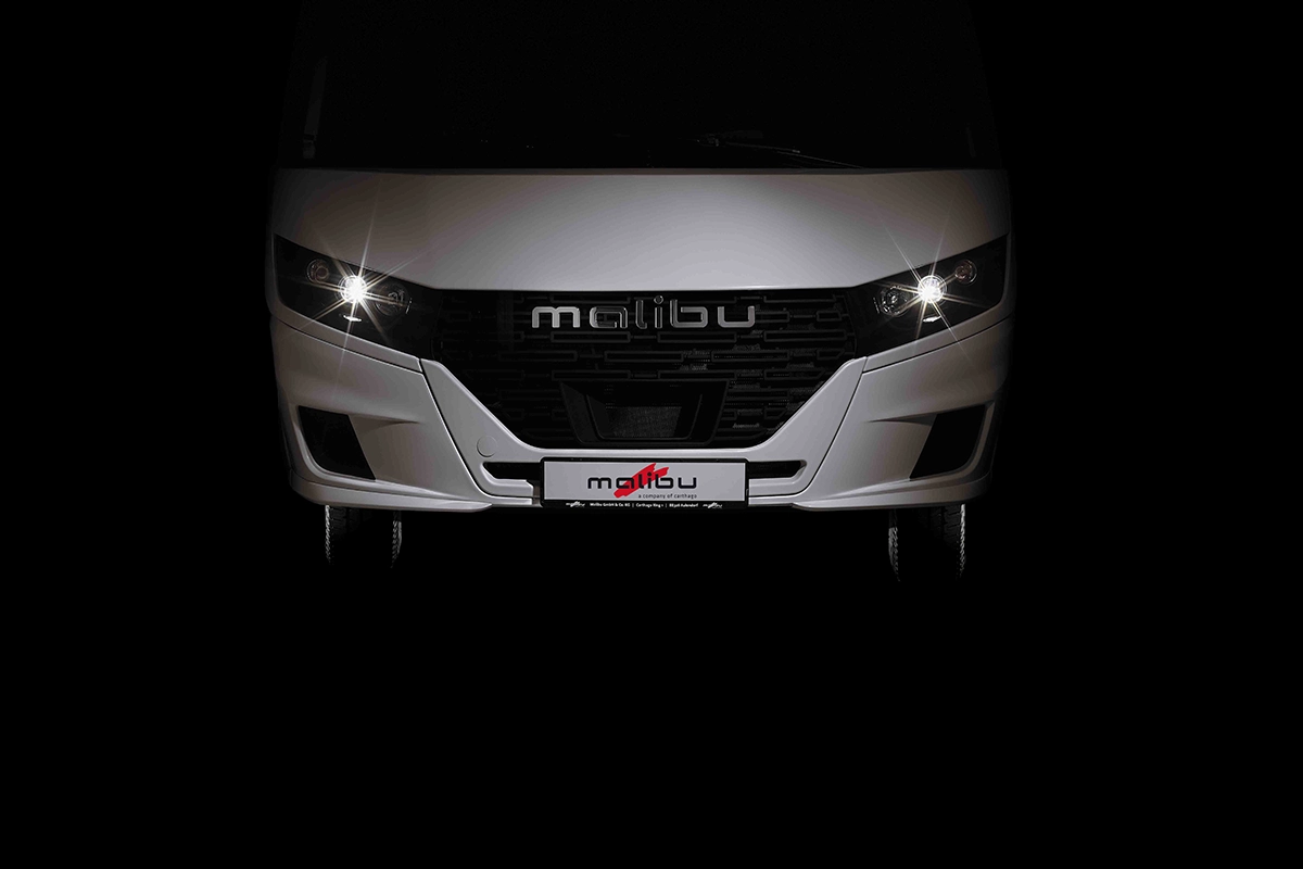 Die Frontansicht eines weißen Malibu-Fahrzeugs ist mit eingeschalteten Scheinwerfern vor einem schwarzen Hintergrund zu sehen. Das Malibu-Logo ist auf dem Kühlergrill und dem Nummernschild zu sehen.