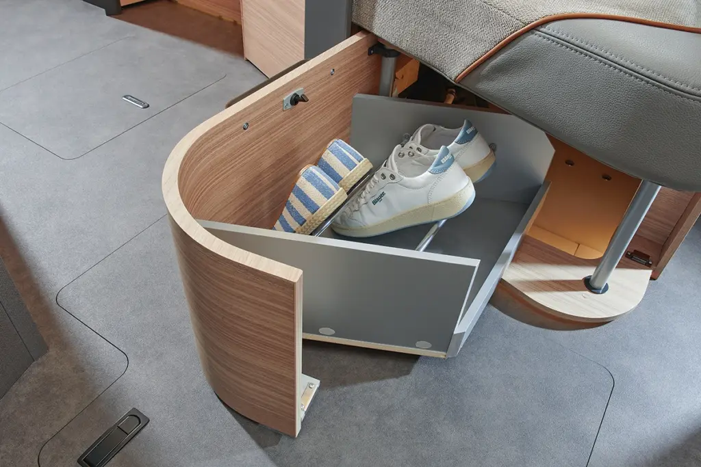 Eine geschwungene Holzschublade unter einem Sitz wird herausgezogen und gibt den Blick auf zwei Paar Schuhe frei - einen lässigen weißen Sneaker und blau gestreifte Espadrilles - in einem modernen, kompakten Wohnraum mit grauem Boden.