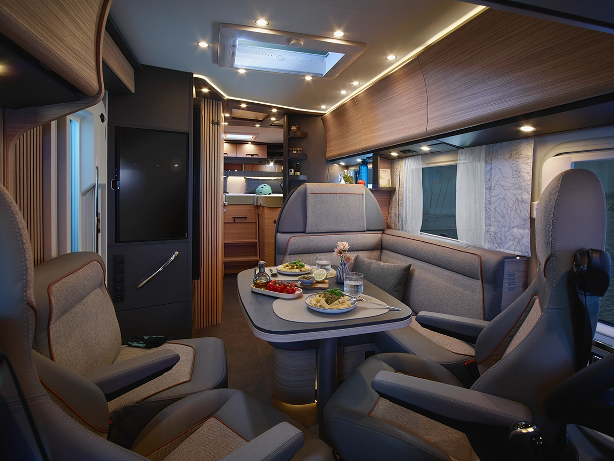 Modernes Wohnmobil-Interieur mit Plüschsitzen, einem mit Lebensmitteln gedeckten Esstisch und einem gut beleuchteten Küchenbereich im Hintergrund. Warme Beleuchtung und Holzakzente schaffen eine gemütliche, einladende Atmosphäre.